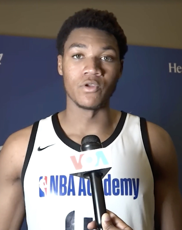 Ein Mann in einem weißen Tanktop spricht bei der NBA All-Star Game 2019 Medienveranstaltung in ein Mikrofon, mit einem Banner und einer Wand im Hintergrund.