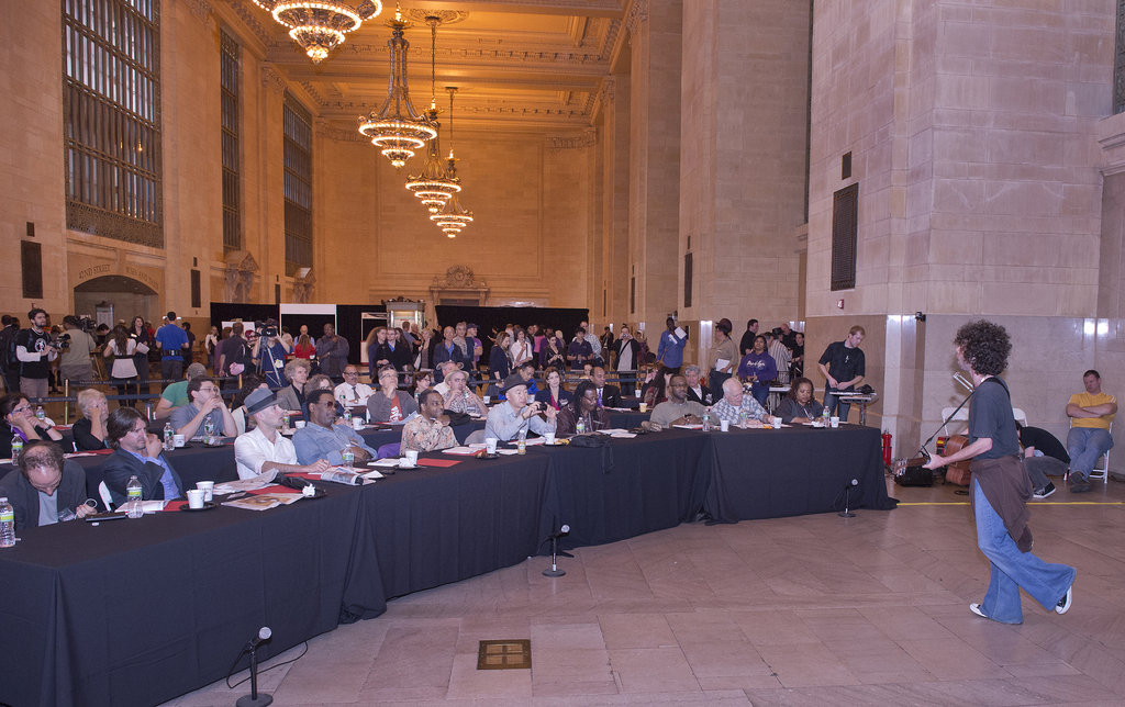 Eine Konzertszene mit einer Menge Menschen, einem Mann, der rechts Gitarre spielt und singt, einer Wand und Fenstern im Hintergrund, Lichtern oben und einem Boden unten.