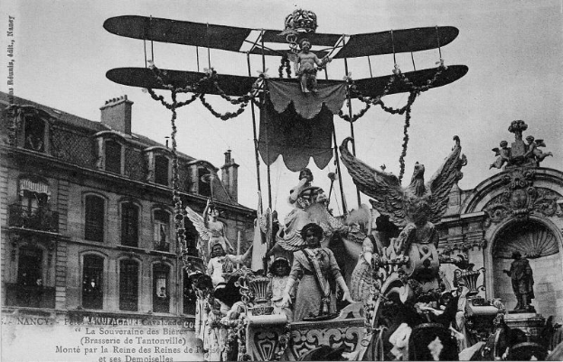 Ein Schwarz-Weiß-Foto eines Umzugswagens mit Statuen und einem Flugzeug, der eine Gruppe von Menschen trägt, mit einem Gebäude und Himmel im Hintergrund und Text unten, der darauf hinweist, dass es ein Foto des Pariser Karnevals ist.