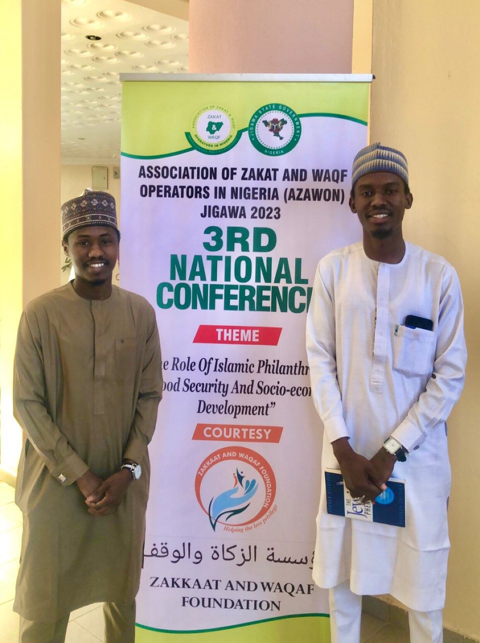 Zwei Männer neben einem "3. Nationalkonferenz"-Plakat mit einer Säule und Deckenleuchten im Hintergrund.