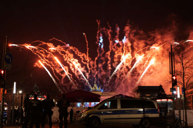 Ein Polizeiwagen steht vor einer Menschenmenge während der Silvesterfeier in Berlin, mit farbenfrohen Feuerwerken am Himmel und beleuchteten Gebäuden und Bäumen im Hintergrund.