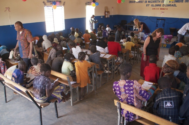 Kinder auf Bänken im Vordergrund mit Erwachsenen in der Nähe, während Stühle, eine Tür, ein Fenster, ein Tisch, Luftballons und Text im Hintergrund zu sehen sind.