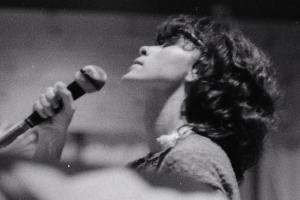 Eine Frau in einem weißen Kleid singt auf der Bühne in ein Mikrofon, ihre Augen geschlossen und ihr Gesichtsausdruck zeigt Emotionen.