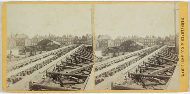 Altes Schwarz-Weiß-Foto einer Bahnschiene mit Gebäuden im Hintergrund, Text 'Merkelbach & Co, Co. Amsterdam' auf der rechten Seite und verstreute hölzerne Gegenstände im Vordergrund.