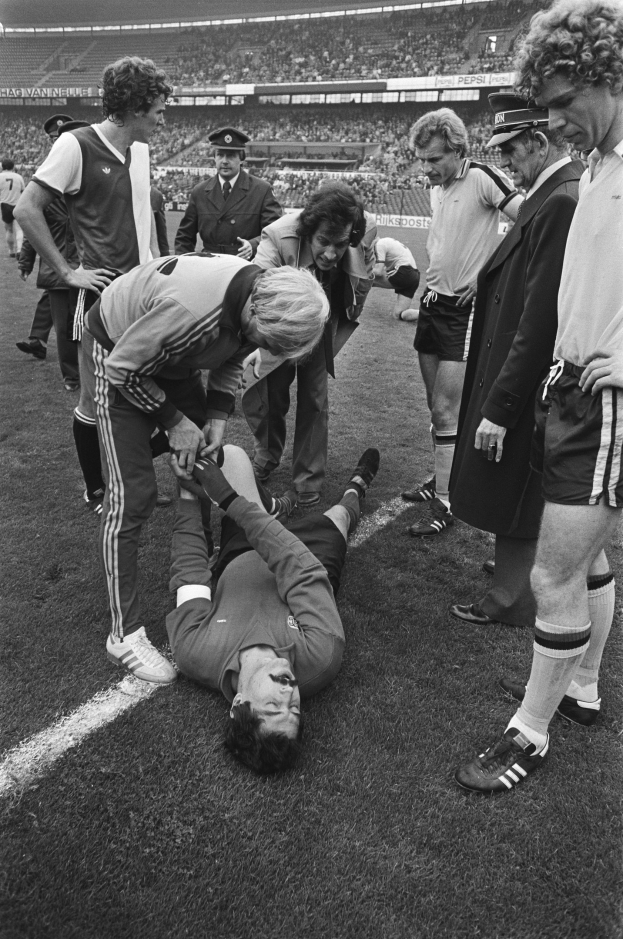 Schwarzes und weißes Foto eines Fußballspielers, der auf dem Boden liegt und von einer Gruppe Männer umgeben ist, mit einer Menschenmenge im Hintergrund eines Stadions.