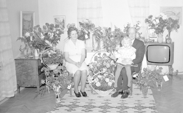 Schwarzes und weißes Foto einer Familie aus drei Personen - einem Mann, einer Frau und einem Kind - auf einem Sofa vor einem Fernseher sitzend, mit Blumenvasen auf den Seitentischen, einem Teppich auf dem Boden, gerahmten Bildern an der Wand und Vorhängen im Hintergrund.