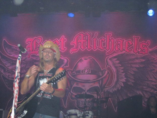 Eine Musikkonzertszene mit einem beleuchteten Banner im Hintergrund, einer Person, die links eine Gitarre spielt und ein Mikrofon vor sich hat, Drogen in der Mitte unten und einer weiteren Person, die in der rechten unteren Ecke sitzt.