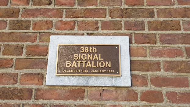 Eine Ziegelwand mit einem rechteckigen Schild, auf dem in fetter, weißer, zentrierter Schrift "38. Signalbataillon" steht.
