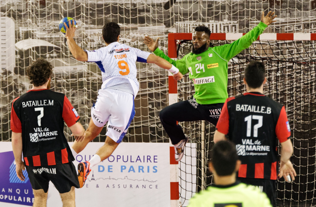 Eine Gruppe von M├Ąnnern, die Handball spielen, mit einem, der springt und einen Ball h├Ąlt, vor einem Tor und einer Tafel mit der Aufschrift "Ligue 1 - Bordeaux vs Marseille."