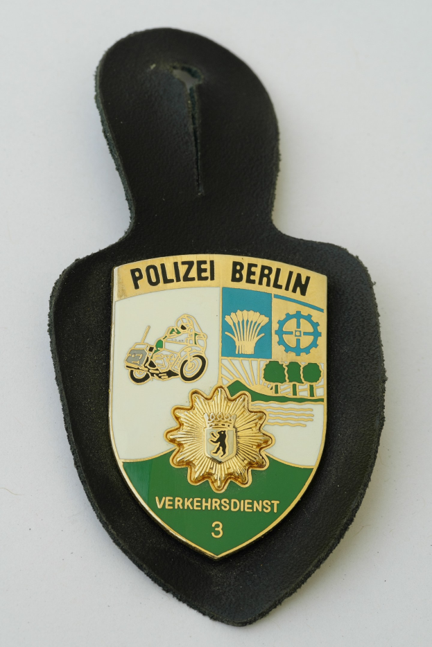 Ein polizeiliches Abzeichen in Form eines Schildes mit der Aufschrift "Polizei Berlin" in schwarzer, fetter Schrift auf einem blauen Hintergrund mit einem weißen Stern und Rahmen, das auf einer Oberfläche platziert ist.