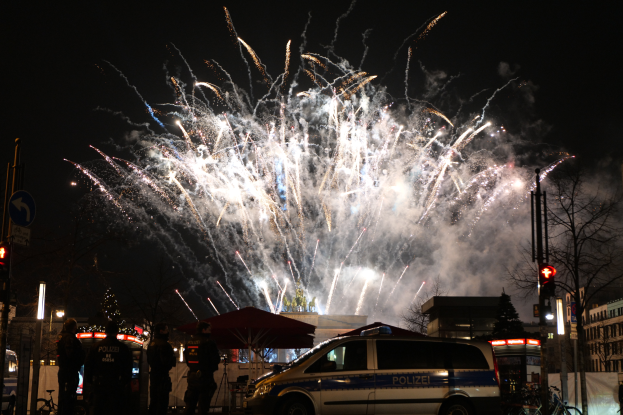 Ein Polizeiwagen steht vor einer Menschenmenge während einer Silvesterfeier in Berlin, mit farbigen Feuerwerken, die den Nachthimmel erhellen.