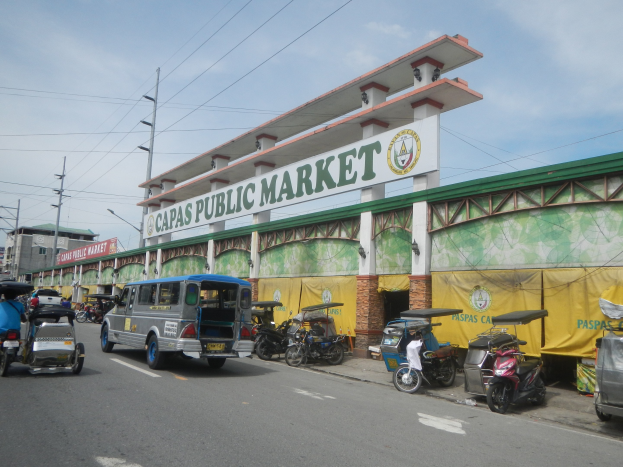 Belebte Stadtstraße mit Autos, Motorrädern und Rikschas vor dem Gebäude "Capas Public Market", Strommasten, Laternenmasten, Gebäuden und bewölktem Himmel.