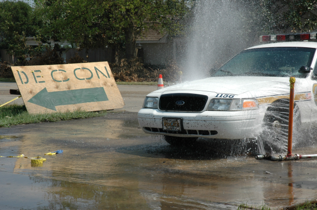 Ein Streifenwagen fährt durch eine Pfütze mit verstreuten Gegenständen, darunter eine Tafel mit Text und ein Verkehrskegel, vor einem Hintergrund aus Bäumen, Gebäuden und einem klaren blauen Himmel.