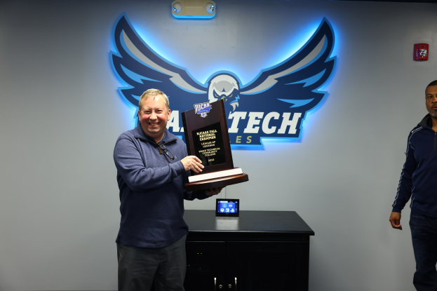 Zwei Männer stehen nebeneinander, einer hält einen Pokal, mit einem Gerät auf dem Tisch vor ihnen und Text mit einem Logo an der Wand dahinter.