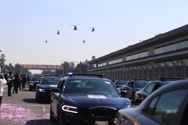 Eine Gruppe von Polizeiwagen vor einem Gebäude geparkt, mit Menschen auf der linken Seite der Straße stehend, Bäumen, Polen, einer Brücke und einem klaren blauen Himmel im Hintergrund und vier Hubschraubern, die oben im Bild fliegen.