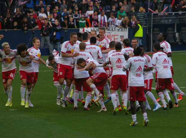 Eine Gruppe von Fußballspielern feiert ein Tor auf einem Rasenfeld, mit Zuschauern in den Rängen, die Kameras und Schilder halten, und Geländern und Stühlen um das Feld herum.