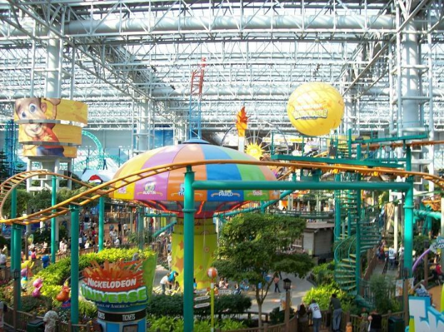 Großes Indoor-Wasserpark mit Achterbahn, umgeben von Menschen, Bäumen, Pflanzen und Deckenstangen mit Lichtern, die eine lebendige Atmosphäre schaffen.