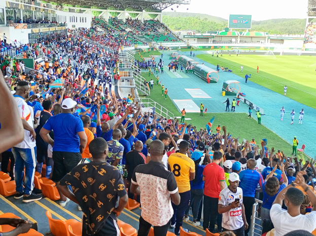 Eine große Menschenmenge versammelt sich vor einem eingezäunten Fußballfeld, wobei einige auf Stühlen sitzen und andere stehen, mit Blick auf Hügel unter einem klaren blauen Himmel während der 2019 African Cup of Nations.