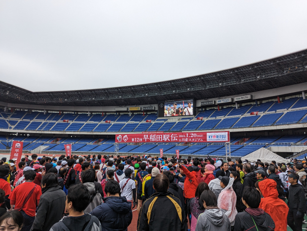 Eine große Menschenmenge steht vor einem Stadion während der Eröffnungszeremonie der Olympischen Spiele in Tokio 2020, mit Bannern, Stühlen und einem Bildschirm im Hintergrund, unter einem sichtbaren Himmel und einem Wasserzeichen in der rechten unteren Ecke.