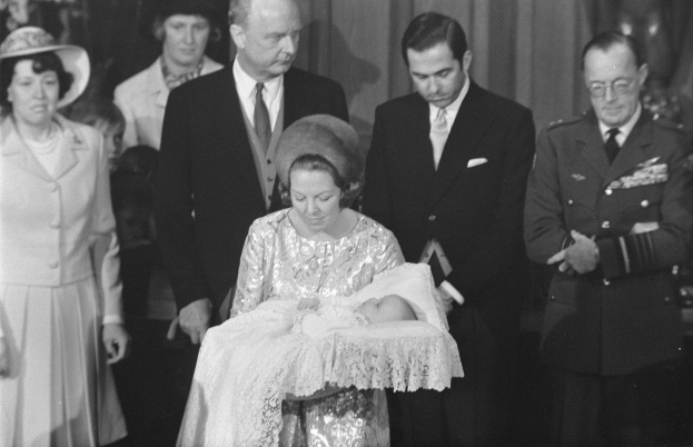Ein Mann und eine Frau sitzen auf einem Stuhl, wobei die Frau ein Baby hält, dahinter ein Fenster mit einem Vorhang und unten ein Text mit der Aufschrift "Die royale Familie".