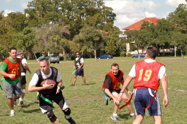 Menschen beim American Football auf einem grünen Feld mit Bäumen, einem Haus und Autos im Hintergrund.