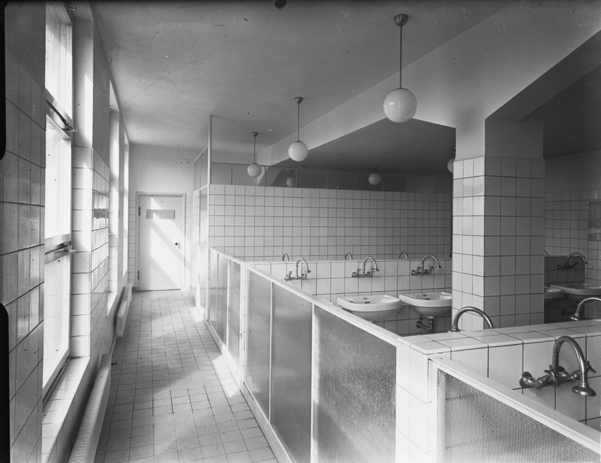 Schwarzes und weißes Foto einer öffentlichen Toilette mit Waschbecken, Wasserhähnen, Deckenleuchten, Wänden, Fenstern und einer Tür.