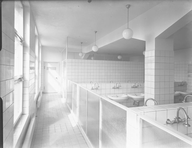 Schwarzes und weißes Foto einer öffentlichen Toilette mit Waschbecken, Wasserhähnen, Deckenleuchten, Wänden, Fenstern und einer Tür.