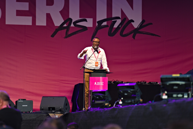 Mann in weißem Hemd und braunen Hosen hält Mikrofon auf Bühne, mit pinkem Banner 'Berlin as Fuck' im Hintergrund und Publikum davor.