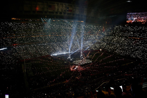 Eine große Menschenmenge in einem hell erleuchteten Stadion bei Nacht schaut auf einen Bildschirm auf der rechten Seite, der die Ergebnisse der Super Bowl XLVII-Halbzeitshow anzeigt.