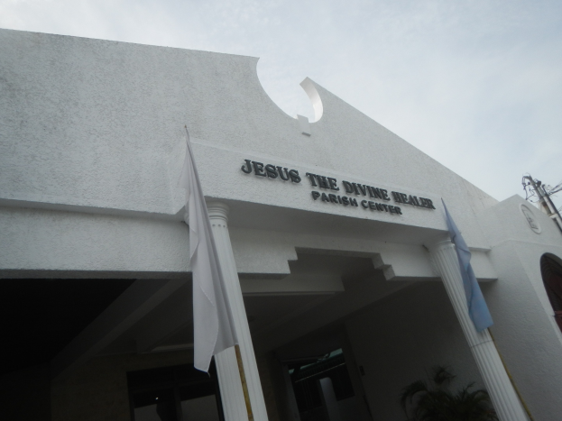 Außenansicht des Jesus der Heilerin Parish Center mit Text an der Wand, Säulen, Fahnen an Stangen, einer Pflanze, einem Strommast mit Drähten und einem bewölkten Himmel als Hintergrund.