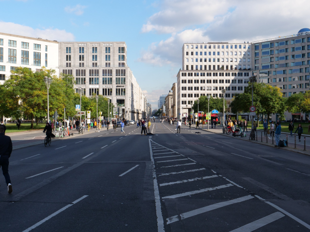 Eine belebte Stadtstraße in Berlin, Deutschland, mit Fußgängern und Radfahrern auf der Straße, hohen Gebäuden, Bäumen, Laternen und Schildern.