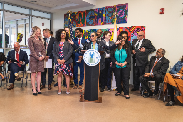 Gruppe von Menschen vor einem Rednerpult mit Mikrofon und einer Tafel mit Text stehen, einige sitzen auf Stühlen und andere stehen, mit Flaggen, Feuerlöscher und Deckenleuchten an der Rückwand.