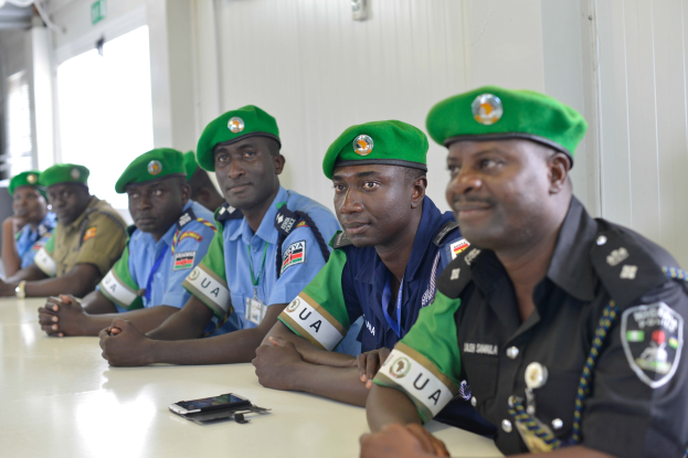 Gruppe von Polizisten, die an einem Tisch während einer Schulung sitzen, mit Mobiltelefonen und anderen Gegenständen auf dem Tisch, in einem Raum mit Fenstern und Deckenbeleuchtung.