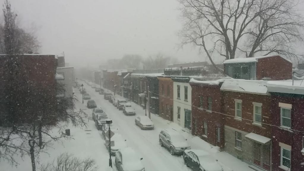 Eine schneebedeckte Straße mit Autos und Gebäuden auf beiden Seiten und Bäumen im Hintergrund.