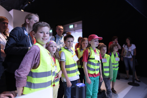 Eine Gruppe von Kindern trägt Sicherheitswesten und Namensschilder und steht auf einem Boden, wobei ein Junge auf der linken Seite lächelt, ein Pfahl auf der rechten Seite steht und ein paar Menschen im Hintergrund zu sehen sind.