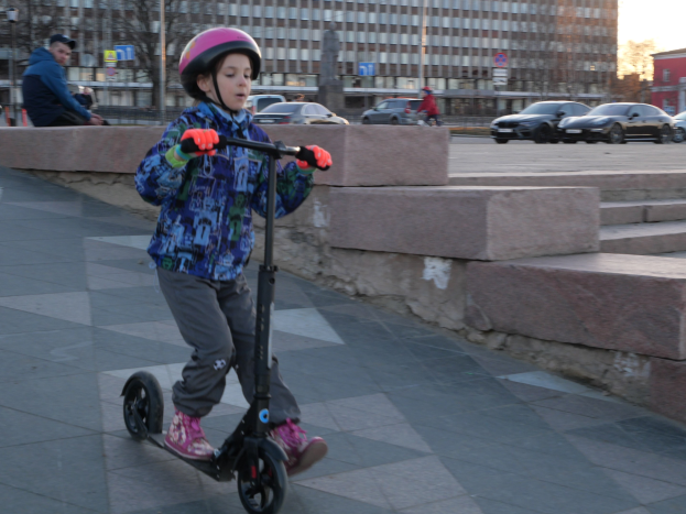 Ein junger Junge in Helm und Handschuhen fährt ein Scooter eine Treppe hinunter auf einem Bürgersteig mit Fahrzeugen, Menschen, Bäumen, Pfählen, Brettern, Gebäuden und einem klaren blauen Himmel im Hintergrund.