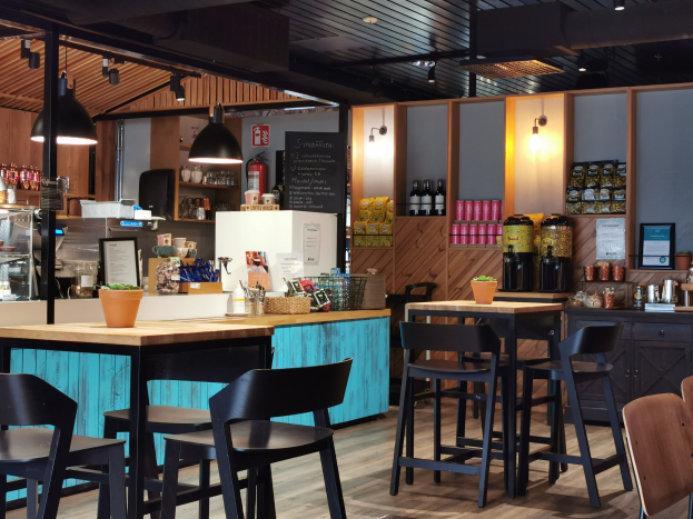 Innenraum eines Restaurants mit Holztischen und -stühlen, einem Topf mit einer Pflanze, einer Schiefertafel, einem Kühlschrank, einem Feuerlöscher, Regalen mit Flaschen, Schränken mit Gegenständen, einem Rahmen mit einem Foto an der Wand und Deckenleuchten.