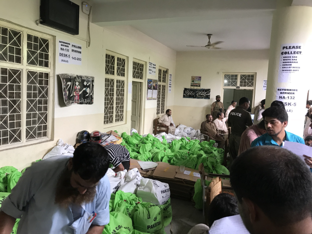 Menschen versammeln sich in einem Raum mit Lebensmittelbeuteln, einige sitzen und andere stehen, halten Papiere, in der Nähe von Fenstern mit Plakaten und einem montierten Fernseher, mit Türen und einer Säule im Hintergrund.