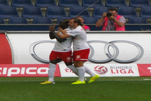 Zwei Fußballspieler feiern ein Tor auf einem Feld umgeben von einer Gruppe von Menschen, mit einer Person, die eine Kamera hält, einem Banner mit Text, einigen Stühlen und einer Wand im Hintergrund.