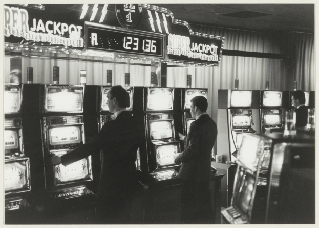 Schwarß-weißes Foto von zwei Männern, die vor Spielautomaten in einem Casino stehen, einer hält ein Glas, mit Vorhängen und einer Texttafel im Hintergrund.