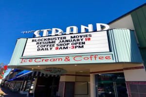 Der Strand-Blockbuster-Movies-Start am 18. Januar Kaffee-Shop-Gebäude mit einem Namensschild, einem Straßenschild, einer Baumgruppe, einigen Topfpflanzen und einem bewölkten Himmel.