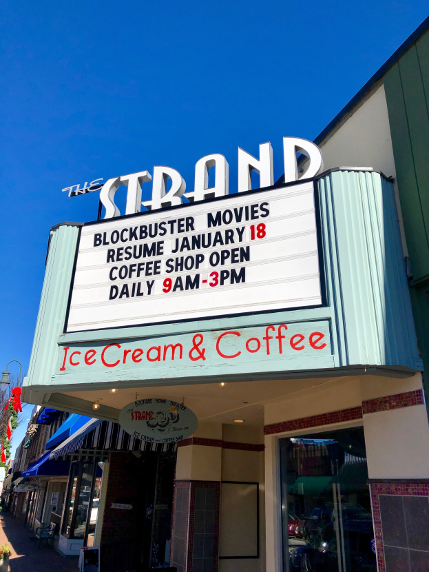 Der Strand-Blockbuster-Movies-Start am 18. Januar Kaffee-Shop-Gebäude mit einem Namensschild, einem Straßenschild, einer Baumgruppe, einigen Topfpflanzen und einem bewölkten Himmel.