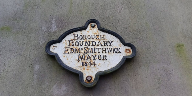 Eine Metalltafel an einem Gebäude mit schwarzem Hintergrund und weißer, fetter, zentrierter Schrift, die "Gemeindegrenze Edm Smithwick Bürgermeister" liest.