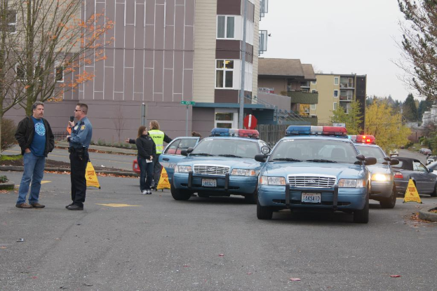 Autos auf einer Straße mit vier Personen in der Nähe, Gebäude mit Fenstern im Hintergrund, Bäume und was aussehen wie Notfallwarndreiecke.