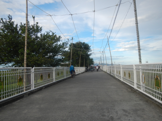 Eine Fußgängerbrücke mit Radfahrern, Geländern, Strommasten, Bäumen und einer bewölkten Himmel