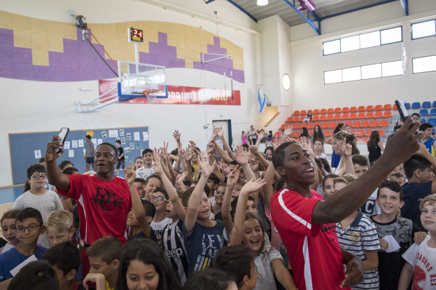 Kinder auf einem Basketballfeld mit Mobiltelefonen, mit einem Tor, einem Basketballkorb, einer Uhr, an die Wand geklebten Papieren, Deckenlampen, Stühlen und Fenstern im Hintergrund.