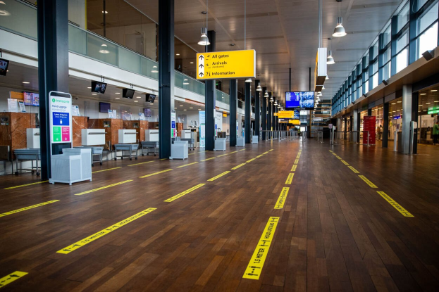 Ein langer Flughafenkorridor mit gelbem Absperrband auf dem Boden, Säulen, Informationsplakate, Bildschirme, Lampen, Glaswände und Fenster, mit einer Person im Hintergrund.