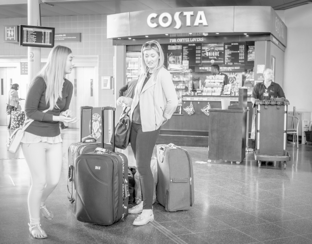 Zwei Frauen mit Gepäck vor einem Kaffeehaus in einer Art Flughafen stehend.