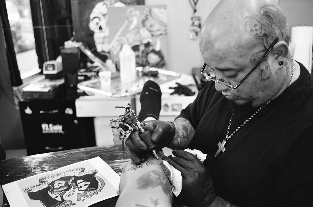 Ein Schwarz-Weiß-Bild eines Mannes in einem schwarzen T-Shirt, mit Brille und einer Kette um den Hals, der ein Bein tätowiert, mit einer Maschine, einem Blatt auf der Oberfläche und einem unscharfen Hintergrund mit einer Flasche, einem Becher und anderen Gegenständen.