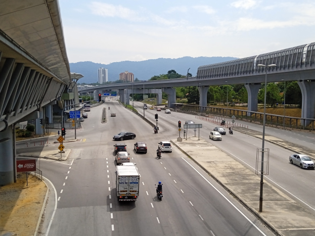Eine vielbefahrene Autobahn mit mehreren Fahrzeugen, neben einer Brücke, mit Straßenlaternen, Verkehrsampeln, Verkehrszeichen, Bäumen, Gebäuden, Hügeln und einem bewölkten Himmel.